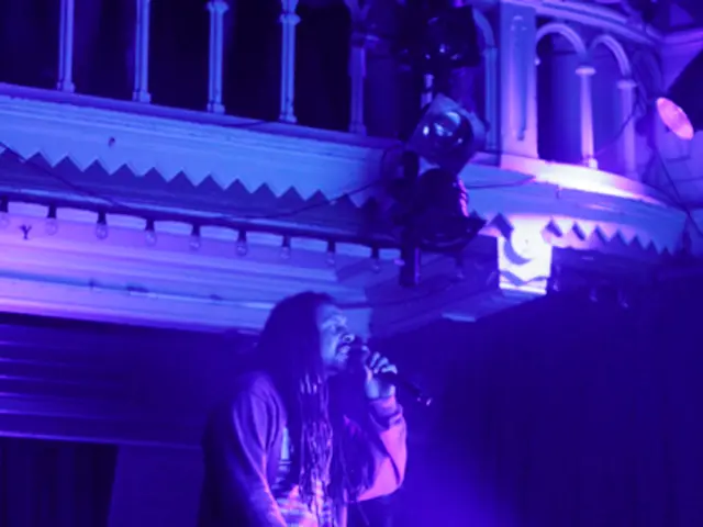 In this image a person is holding a mic. I can see focusing lights, laptops, curtains, railing,...