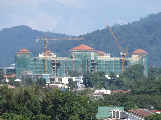 In this image there are few buildings, houses, trees, cranes, mountains covered with trees and some...