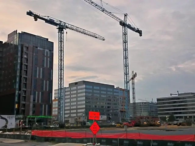 In this image we can see buildings, sky with clouds, construction cranes, polythene cover, sign...